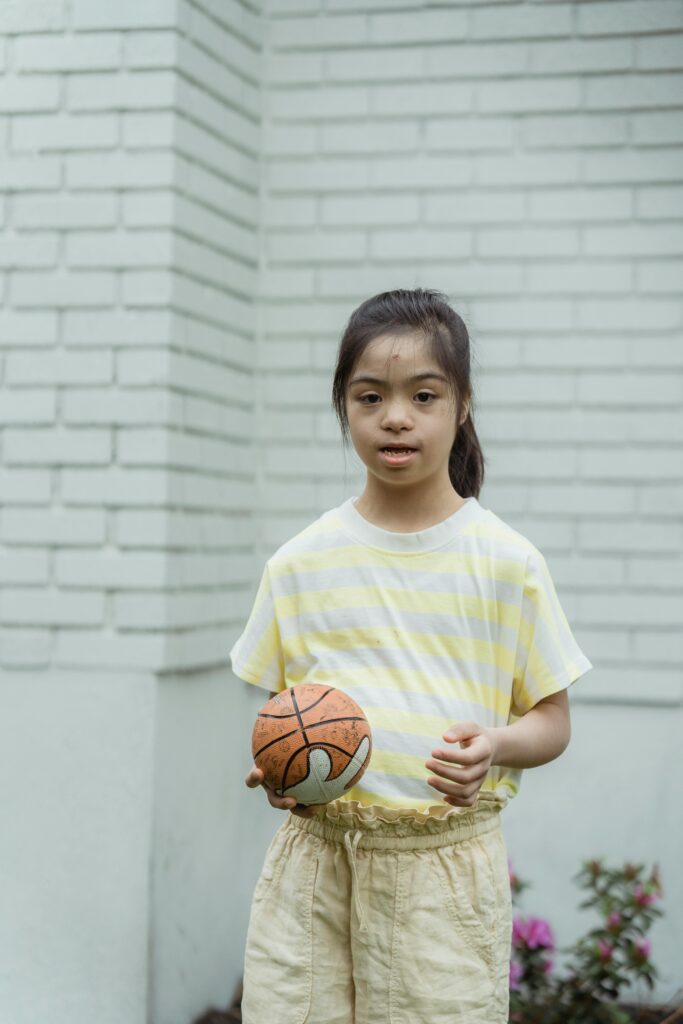 A young girl with Down syndrome holding a basketball outdoors. Captured in soft lighting.